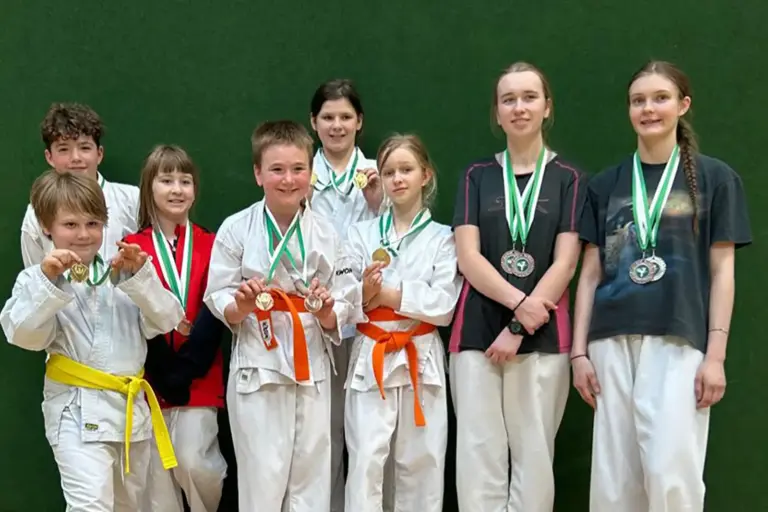 Gruppe von Kindern und Jugendlichen in Karateanzügen und Sportkleidung steht vor einer grünen Wand und zeigt stolz ihre Medaillen. Einige tragen farbige Gürtel, darunter gelb und orange, und halten die Auszeichnungen in die Kamera.