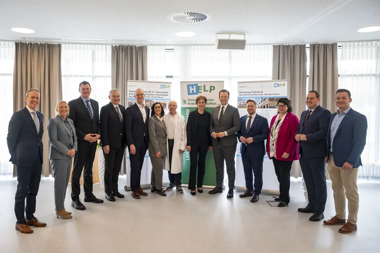 Eine Gruppe von Vertreterinnen und Vertretern aus Politik und Gesundheitswesen steht bei einer Präsentation vor Roll-ups im Innenraum. Die Personen sind formell gekleidet, in der Mitte steht auch medizinisches Personal, im Hintergrund informieren Plakate über Projekte im Krankenhaus.