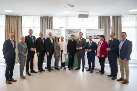 Eine Gruppe von Vertreterinnen und Vertretern aus Politik und Gesundheitswesen steht bei einer Präsentation vor Roll-ups im Innenraum. Die Personen sind formell gekleidet, in der Mitte steht auch medizinisches Personal, im Hintergrund informieren Plakate über Projekte im Krankenhaus.