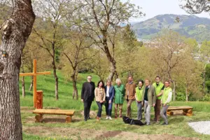 Eine Gruppe von Personen steht neben einem Holzkreuz und rustikalen Holzbänken auf einer Wiese.