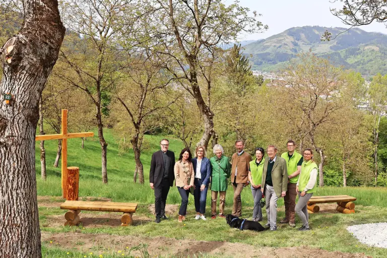 Eine Gruppe von Personen steht neben einem Holzkreuz und rustikalen Holzbänken auf einer Wiese.