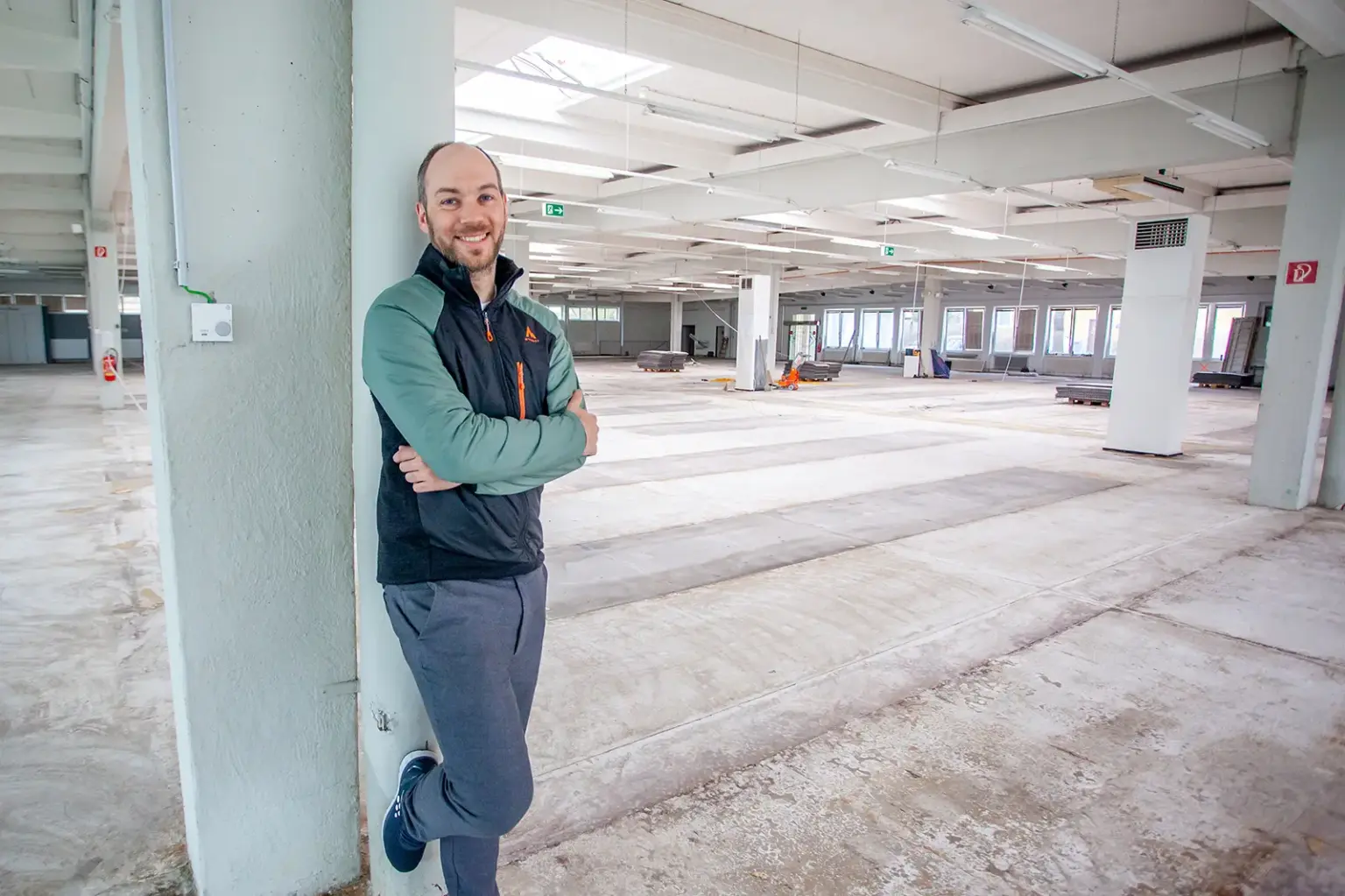 Ein Mann steht an einer Säule in in einer leeren Gewerbehalle, im Hintergrund Material und Geräte für eine Bodensanierung