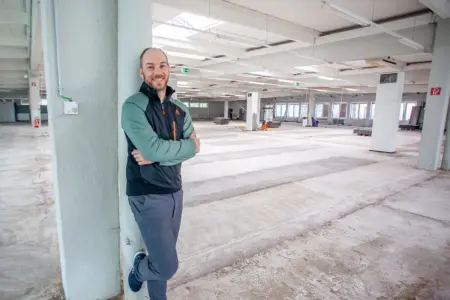 Ein Mann steht an einer Säule in in einer leeren Gewerbehalle, im Hintergrund Material und Geräte für eine Bodensanierung