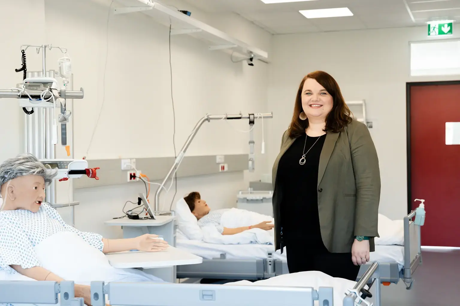 Eine Frau steht in einem modernen Pflege- oder Schulungsraum neben zwei Krankenbetten. In den Betten liegen Übungspuppen, medizinische Geräte sind an den Wänden montiert, die Frau blickt lächelnd in die Kamera.