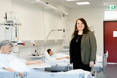 Eine Frau steht in einem modernen Pflege- oder Schulungsraum neben zwei Krankenbetten. In den Betten liegen Übungspuppen, medizinische Geräte sind an den Wänden montiert, die Frau blickt lächelnd in die Kamera.