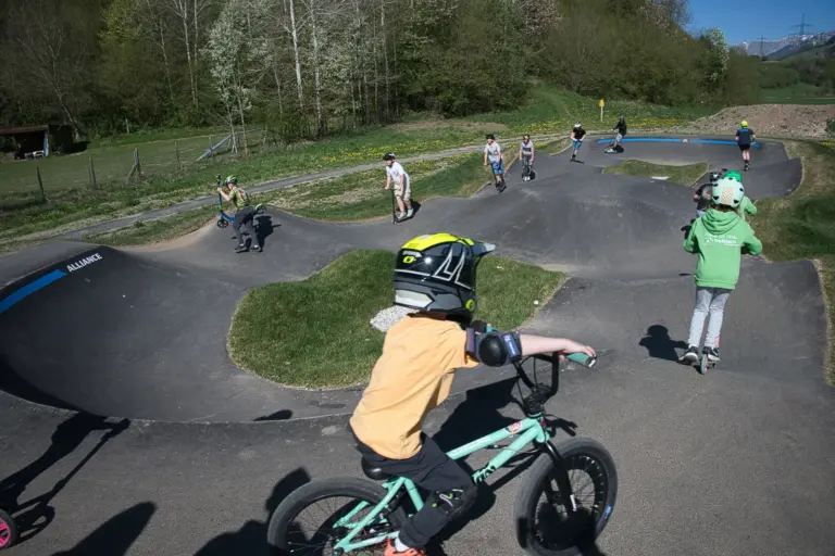 Kinder mit Helmen fahren mit Fahrrädern und Rollern auf einer asphaltierten Buckelbahn
