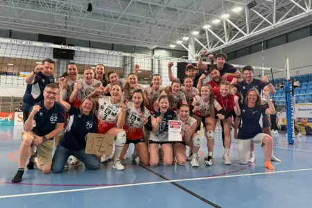 Jubelndes Volleyballteam mit Trainerstab in einer Sporthalle: Die Spielerinnen knien und stehen dicht beisammen vor dem Netz, einige halten Medaillen und eine Urkunde in die Kamera, alle reißen lachend die Arme hoch. Im Hintergrund sind Tribüne und Hallenkonstruktion sichtbar.