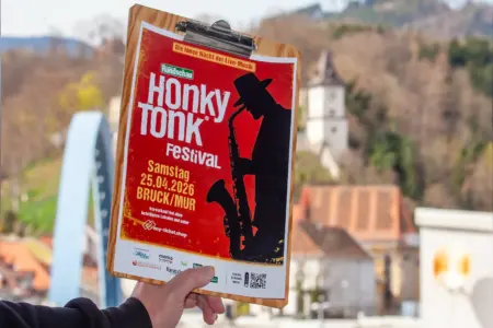 Eine Hand hält ein Klemmbrett mit einem roten Plakat für das „Honky Tonk Festival“. Darauf stehen Termin und Ort (25. April 2026 in Bruck an der Mur), im Hintergrund sind unscharf Häuser, eine Brücke und ein Turm zu sehen.