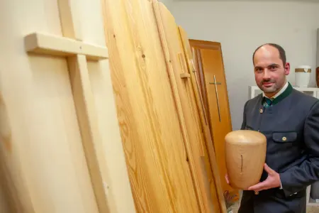 Ein Mann im Trachtenanzug mit einer Urne in Händen, daneben vier Särge mit Kreuzen aus Holz und Metall darauf