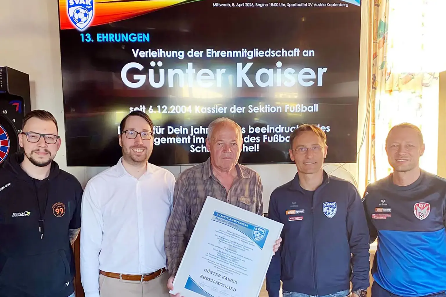 Fünf Männer stehen nebeneinander in einem Vereinsraum, in der Mitte hält ein älterer Mann eine Urkunde in die Kamera. Auf einem Bildschirm im Hintergrund ist die Verleihung der Ehrenmitgliedschaft an „Günter Kaiser“ zu lesen.