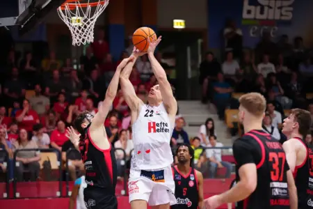 Ein Basketballspieler im weißen Trikot springt unter dem Korb zum Wurf hoch, während ein Gegenspieler im schwarzen Trikot ihn zu blocken versucht; weitere Spieler und Zuschauer sind im Hintergrund zu sehen.