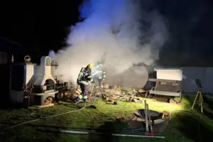 Zwei Feuerwehrleute löschen nachts einen Brand in einem Garten. Zwischen Holzresten und dichter Rauchentwicklung sind sie mit Atemschutz und Schlauch im Einsatz, während nebenan ein gemauerter Grill und Gartenmöbel stehen.