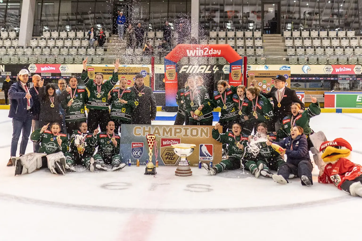 Eine Eishockeymannschaft feiert auf dem Eis den Titelgewinn. Die Spielerinnen tragen Medaillen, sprühen mit Getränken und jubeln rund um einen Pokal, vor einem „Champion“-Schild und einer Eventkulisse in der Halle.