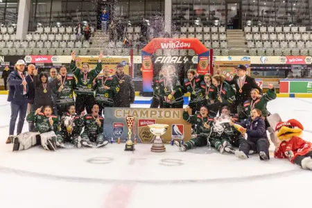 Eine Eishockeymannschaft feiert auf dem Eis den Titelgewinn. Die Spielerinnen tragen Medaillen, sprühen mit Getränken und jubeln rund um einen Pokal, vor einem „Champion“-Schild und einer Eventkulisse in der Halle.