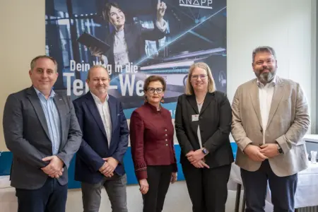 Fünf erwachsene Personen in Businesskleidung stehen nebeneinander in einem Innenraum und blicken in die Kamera. Im Hintergrund hängt ein großes Werbeplakat mit einer Frau, die ein Tablet hält und nach oben zeigt, sowie dem Schriftzug „Dein Weg in die Tech-Welt“.