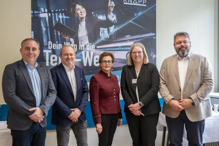 Fünf erwachsene Personen in Businesskleidung stehen nebeneinander in einem Innenraum und blicken in die Kamera. Im Hintergrund hängt ein großes Werbeplakat mit einer Frau, die ein Tablet hält und nach oben zeigt, sowie dem Schriftzug „Dein Weg in die Tech-Welt“.