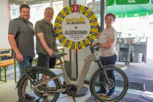 Zwei Männer und eine Frau stehen mit einem E-Bike vor einem Glücksrad der Firma EMB.