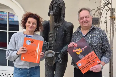 Ein Mann und eine Frau stehen mit einem Plakat und einer Zeitschrift neben der Statue eines Musikers