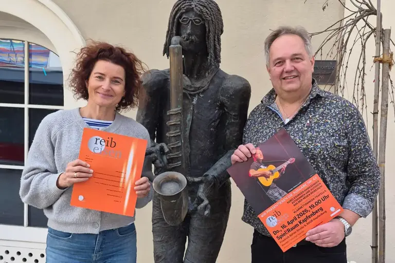 Ein Mann und eine Frau stehen mit einem Plakat und einer Zeitschrift neben der Statue eines Musikers