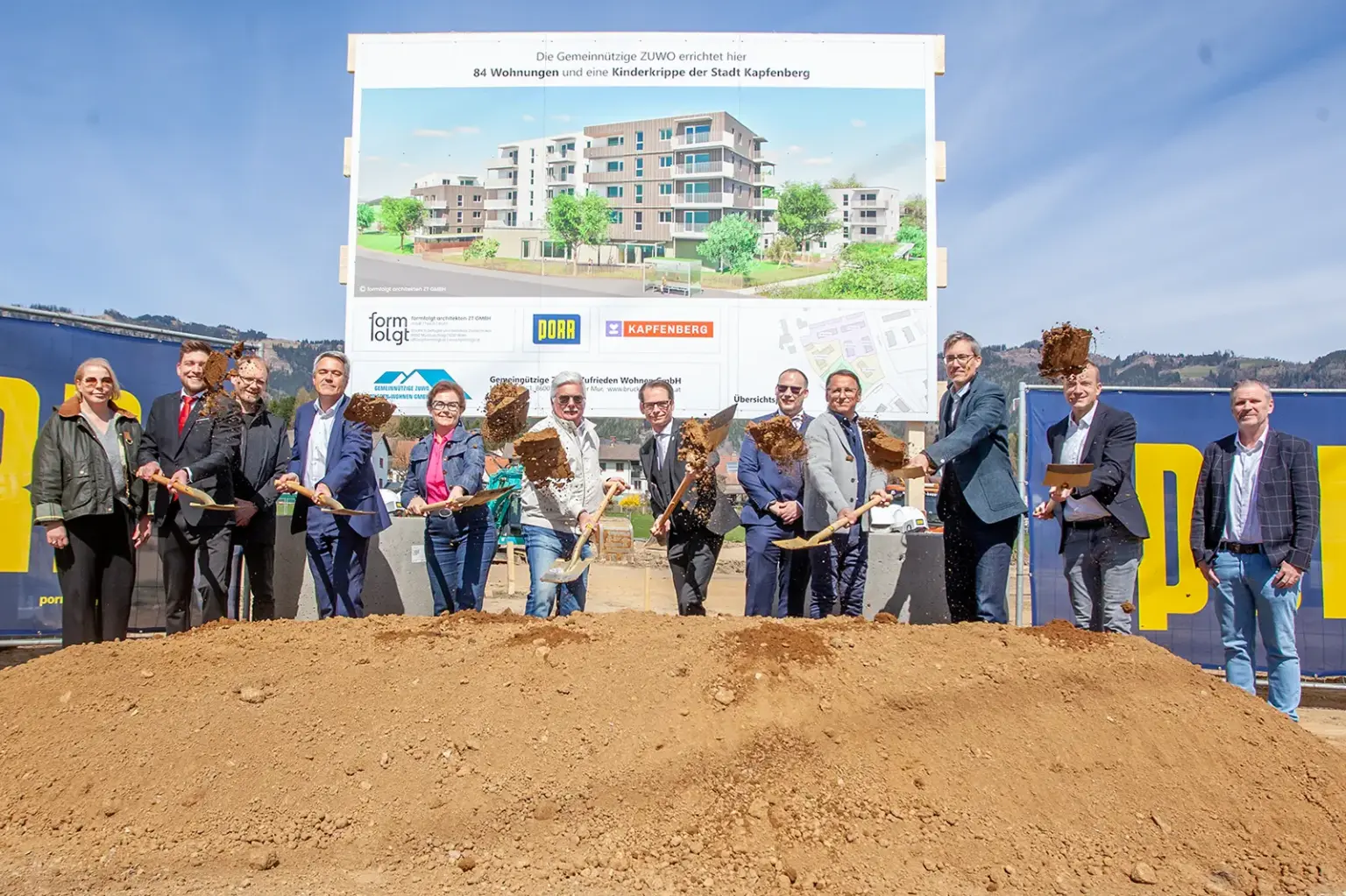 Zwölf Personen bei einer Spatenstichfeier, acht werfen mit Schaufeln lose Erde in Richtung Kamera. Im Hintergrund eine Plakatwand, darauf eine Abbildung von Häusern, teils vier-, teils fünfgeschoßig