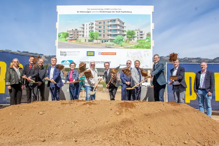 Zwölf Personen bei einer Spatenstichfeier, acht werfen mit Schaufeln lose Erde in Richtung Kamera. Im Hintergrund eine Plakatwand, darauf eine Abbildung von Häusern, teils vier-, teils fünfgeschoßig