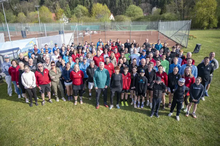 Große Gruppe von Teilnehmerinnen und Teilnehmern eines Tennisevents posiert gemeinsam vor Tennisplätzen im Freien für ein Gruppenfoto.