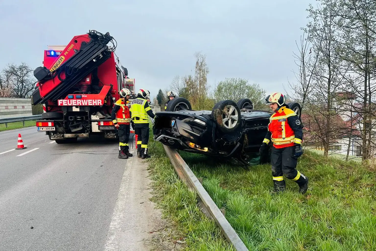 Einsatzkräfte der Feuerwehr sichern einen Verkehrsunfall auf einer Straße. Ein Auto liegt auf dem Dach neben der Leitplanke, während Feuerwehrleute in Schutzkleidung den Einsatzort betreuen, daneben steht ein Feuerwehrfahrzeug.