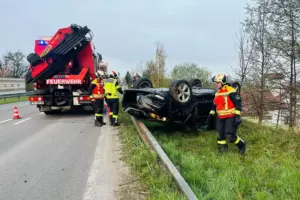 Einsatzkräfte der Feuerwehr sichern einen Verkehrsunfall auf einer Straße. Ein Auto liegt auf dem Dach neben der Leitplanke, während Feuerwehrleute in Schutzkleidung den Einsatzort betreuen, daneben steht ein Feuerwehrfahrzeug.
