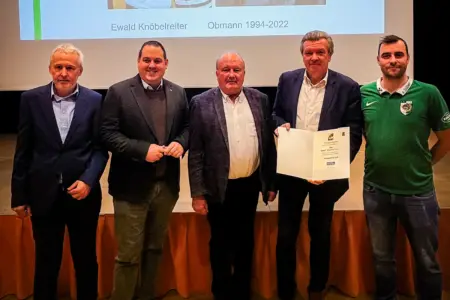 Fünf Männer stehen nebeneinander auf einer Bühne, einer von ihnen hält eine aufgeschlagene Urkunde in die Kamera. Im Hintergrund ist eine Präsentationsfolie mit einem Namen und einer Funktionsbezeichnung zu sehen.