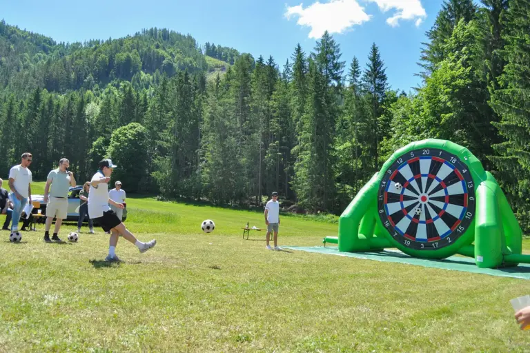Mehrere Personen spielen auf einer Wiese Fußball-Darts. Ein Teilnehmer schießt einen Ball auf eine große aufblasbare Dartscheibe, während andere zuschauen, im Hintergrund Wald und Berge.