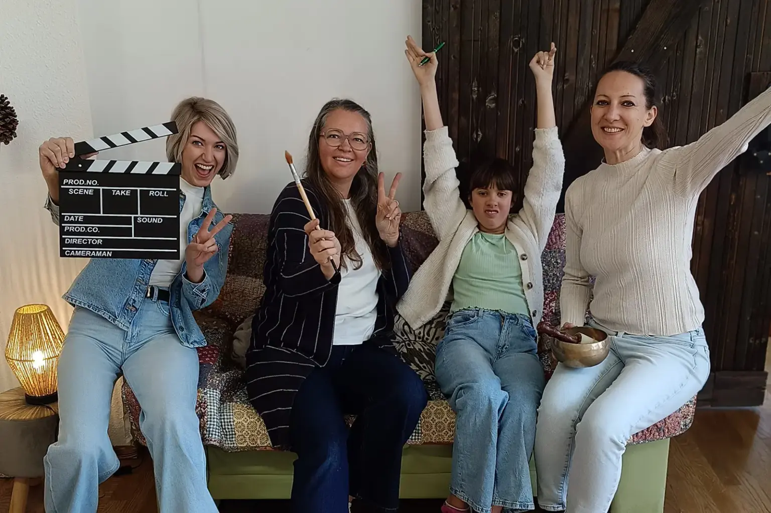 Vier Personen sitzen auf einem Sofa in einem Wohnraum und posieren fröhlich für die Kamera. Eine Frau hält eine Filmklappe, eine andere einen Pinsel, ein Kind hebt die Arme, und alle zeigen eine lockere, kreative Stimmung.