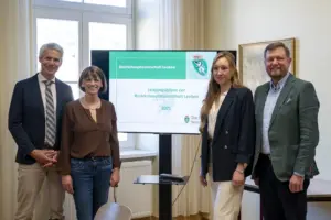 Vier Personen stehen in einem Büro neben einem Bildschirm, auf dem „Leistungsbilanz der Bezirkshauptmannschaft Leoben 2025“ zu lesen ist. Sie blicken in die Kamera, im Hintergrund sind Fenster und ein Besprechungsraum zu sehen.