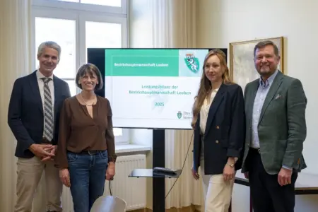 Vier Personen stehen in einem Büro neben einem Bildschirm, auf dem „Leistungsbilanz der Bezirkshauptmannschaft Leoben 2025“ zu lesen ist. Sie blicken in die Kamera, im Hintergrund sind Fenster und ein Besprechungsraum zu sehen.