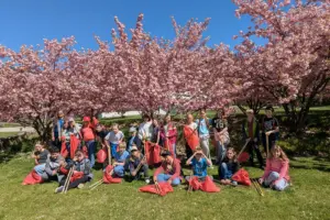 Volksschüler sitzen und stehen auf einer Wiese. Sie halten Säcke und lange Zangen zum Müllsammeln in den Händen.