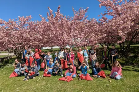 Volksschüler sitzen und stehen auf einer Wiese. Sie halten Säcke und lange Zangen zum Müllsammeln in den Händen.