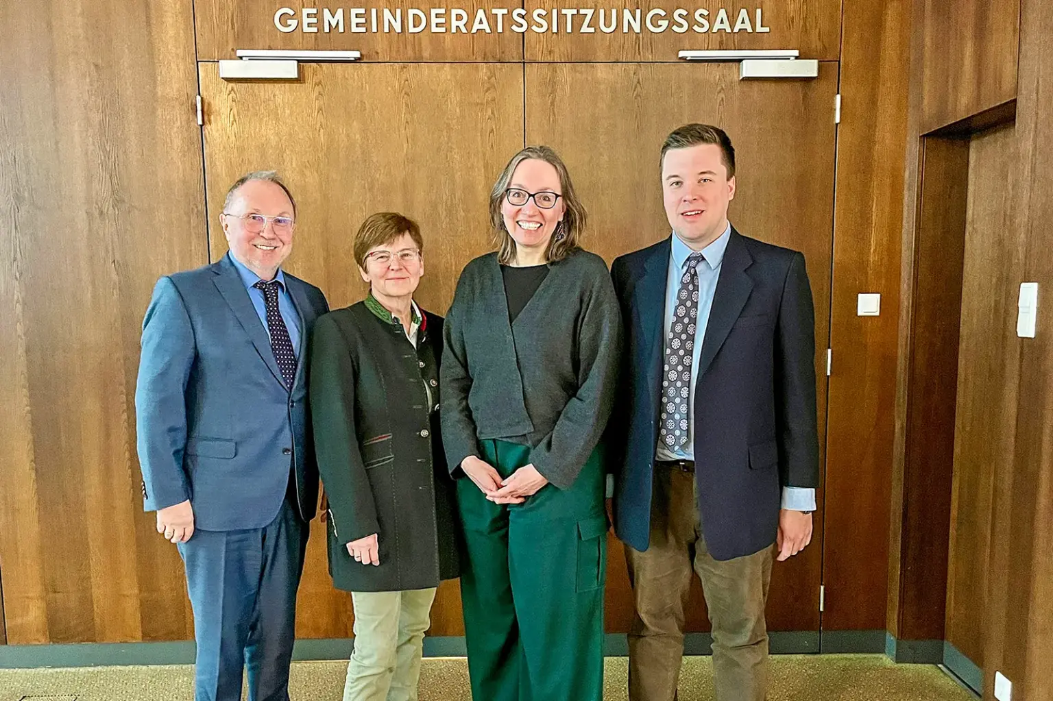 Zwei Frauen und zwei Männer in förmlicher Kleidung vor einer mit Holz vertäfelten Wand, über einer breiten zweiflügeligen Tür die Aufschrift "Gemeinderatssitzungssaal"