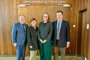 Zwei Frauen und zwei Männer in förmlicher Kleidung vor einer mit Holz vertäfelten Wand, über einer breiten zweiflügeligen Tür die Aufschrift "Gemeinderatssitzungssaal"