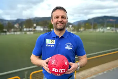 Ein Mann im blauen Poloshirt mit Vereinslogo steht am Rand eines Sportplatzes und hält einen roten Handball in beiden Händen. Er blickt lächelnd in die Kamera, im Hintergrund sind ein Spielfeld und Berge zu sehen.
