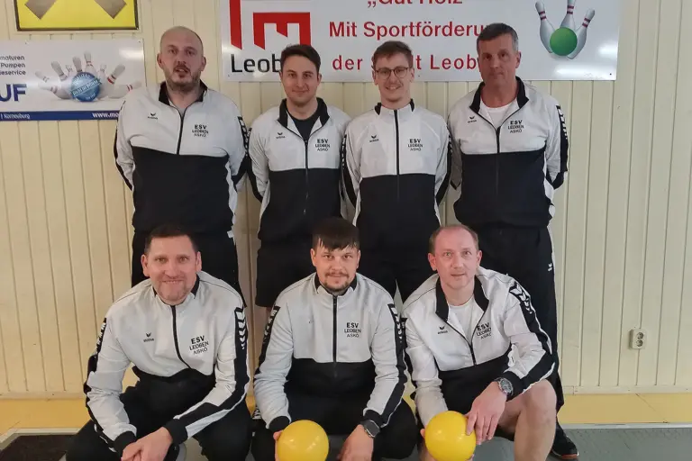 Sieben Männer des ESV Leoben posieren in einheitlichen Trainingsanzügen in einer Kegelhalle; drei knien vorne mit gelben Kugeln, vier stehen dahinter vor einer Wand mit Vereins- und Sponsorenplakaten.