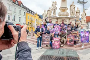 Zwölf Personen mit Musikinstrumenten und bunten Plakaten posieren vor einem Denkmal, am Bildrand im Vordergrund ein Mann, der durch den Sucher einer Fotokamera schaut