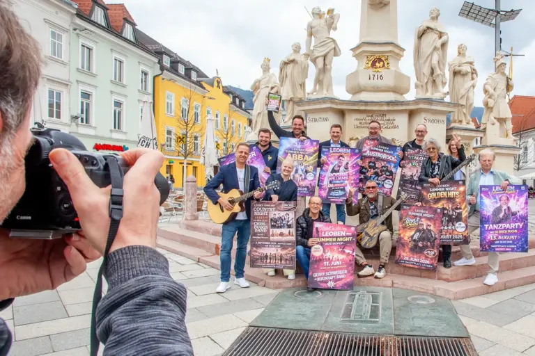 Zwölf Personen mit Musikinstrumenten und bunten Plakaten posieren vor einem Denkmal, am Bildrand im Vordergrund ein Mann, der durch den Sucher einer Fotokamera schaut