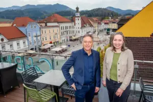 Ein Mann und eine Frau stehen auf einer Terrasse, im Hintergrund ein innerstädtischer Platz