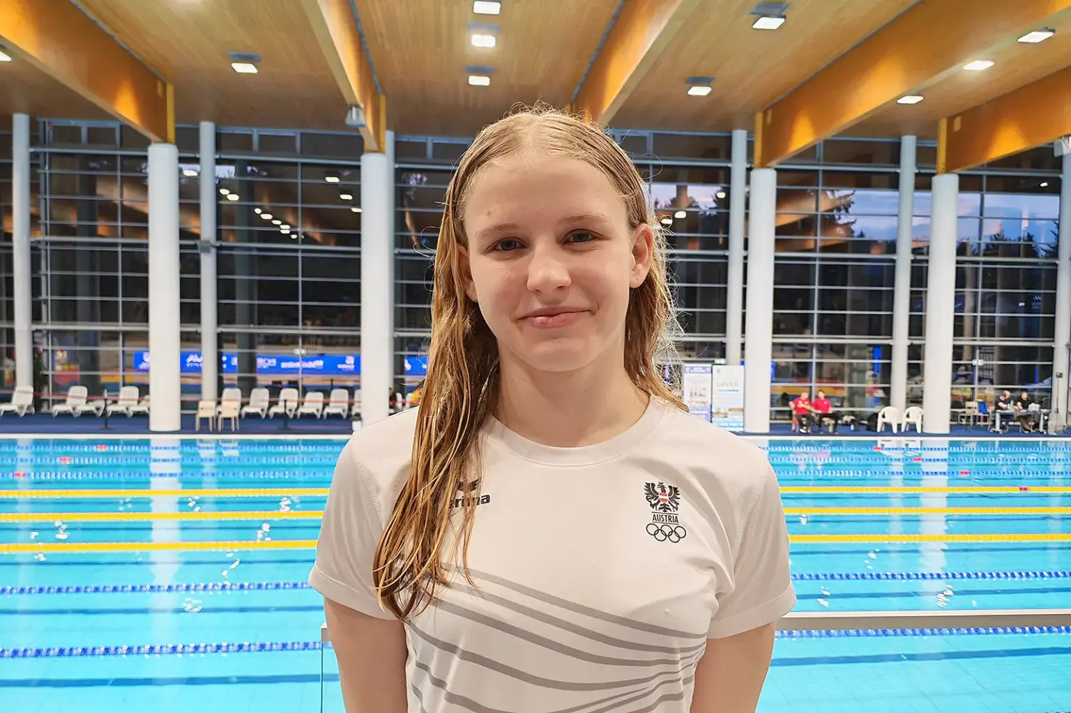 Schnelle Zeiten von Schwimmerin Lilli Paier Die österreichische Schwimmerin Lilli Paier steht am Beckenrand eines Hallenbads in Sopron und blickt in die Kamera; hinter ihr sind mehrere Schwimmbahnen und Sitzreihen zu sehen, ihre Haare sind noch nass vom Training oder Wettkampf.