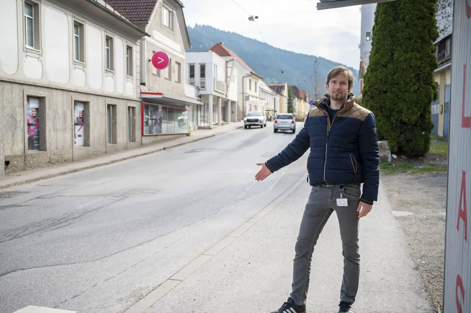 Mürzzuschlag: Baustart für die Sanierung der Fernwärmeleitung Ein Mann steht am Gehsteig einer Ortsstraße und zeigt mit ausgestrecktem Arm auf die Fahrbahn. Im Hintergrund sind Häuser, parkende Autos und bewaldete Hügel zu sehen.