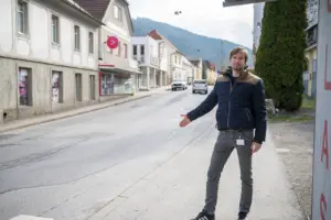 Ein Mann steht am Gehsteig einer Ortsstraße und zeigt mit ausgestrecktem Arm auf die Fahrbahn. Im Hintergrund sind Häuser, parkende Autos und bewaldete Hügel zu sehen.