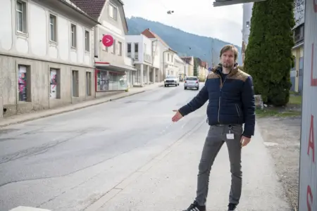 Ein Mann steht am Gehsteig einer Ortsstraße und zeigt mit ausgestrecktem Arm auf die Fahrbahn. Im Hintergrund sind Häuser, parkende Autos und bewaldete Hügel zu sehen.