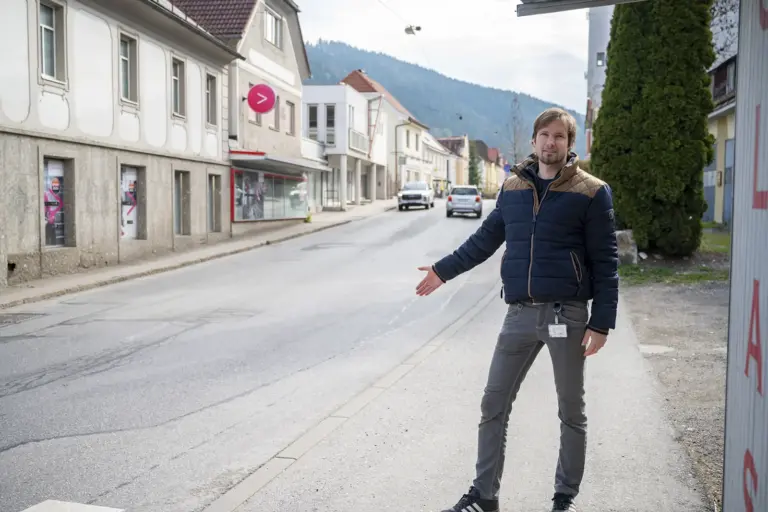 Ein Mann steht am Gehsteig einer Ortsstraße und zeigt mit ausgestrecktem Arm auf die Fahrbahn. Im Hintergrund sind Häuser, parkende Autos und bewaldete Hügel zu sehen.
