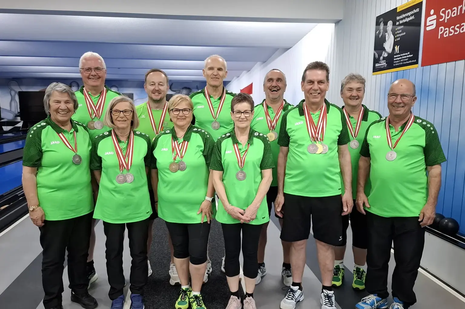 Gruppe von Frauen und Männern in grünen Sportshirts steht in einer Kegelbahn und präsentiert ihre Medaillen. Die Teilnehmer lächeln in die Kamera, im Hintergrund sind Bahnen und Vereinsplakate zu sehen.
