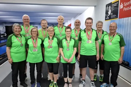 Gruppe von Frauen und Männern in grünen Sportshirts steht in einer Kegelbahn und präsentiert ihre Medaillen. Die Teilnehmer lächeln in die Kamera, im Hintergrund sind Bahnen und Vereinsplakate zu sehen.