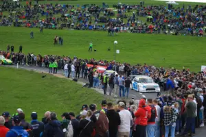 Zahlreiche Zuschauerinnen und Zuschauer säumen eine Straße bei einem Bergrennen. Entlang der Strecke stehen dicht gedrängt Menschen, während Rennfahrzeuge vorbeifahren, im Hintergrund sitzen weitere Besucher auf einer Wiese.